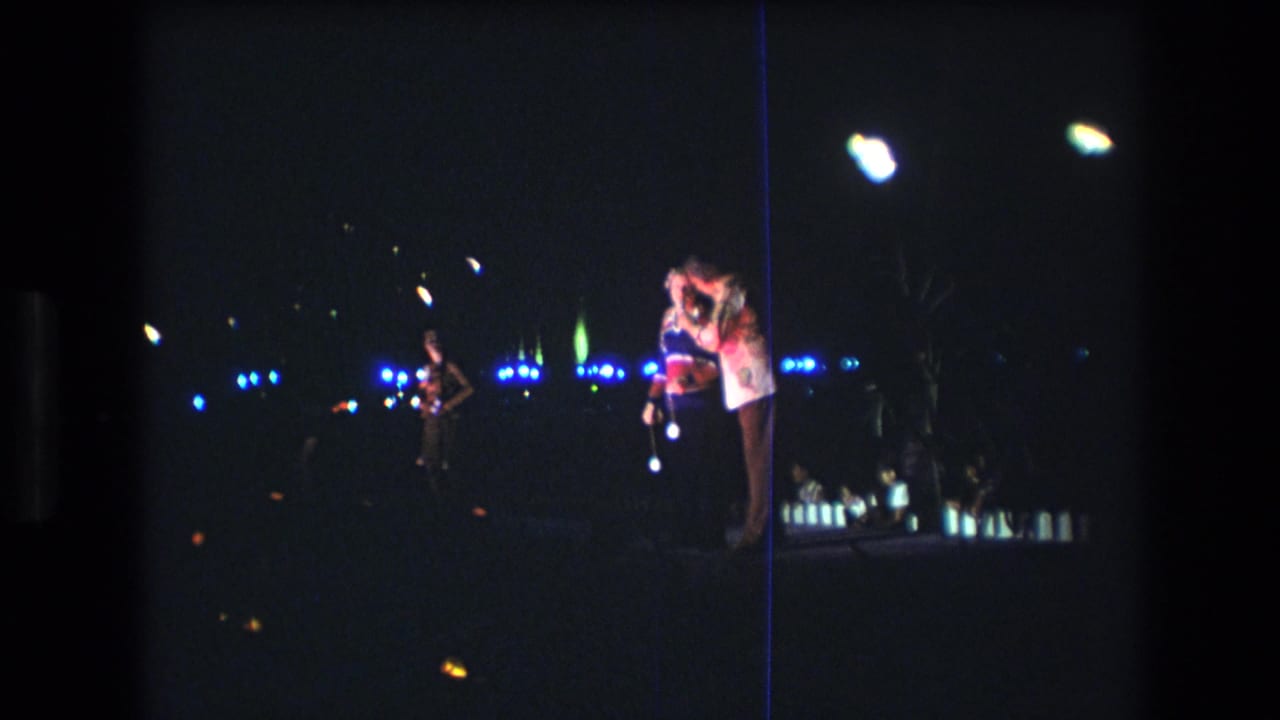 1977 Honolulu home-movie frame showing a man and woman standing close together at night near the coast, with blue background lights and film flare visible.