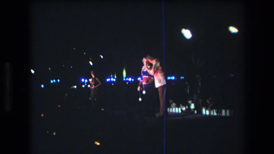 1977 Honolulu home-movie frame showing a man and woman standing close together at night near the coast, with blue background lights and film flare visible.