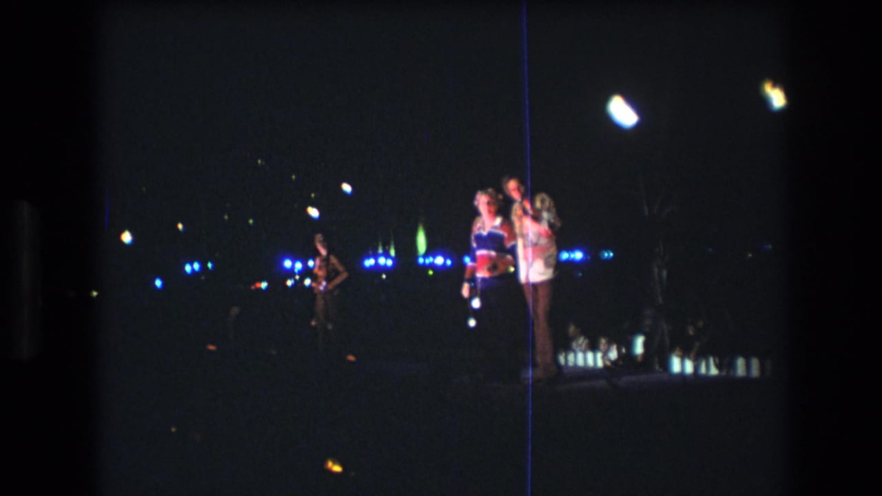 1977 Honolulu 8mm frame of a man in a patterned shirt beside a woman in a colorful top, with distant blue lights and dark coastal surroundings.