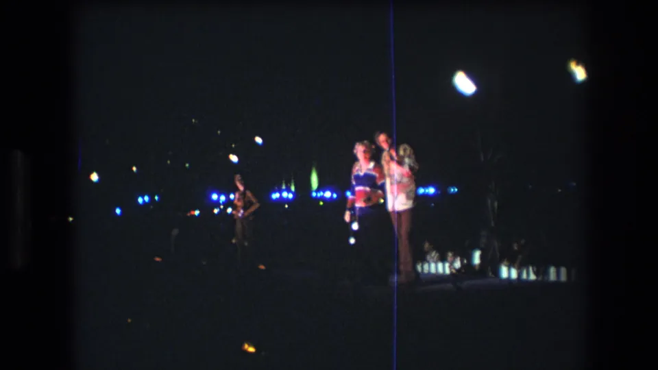 1977 Honolulu 8mm frame of a man in a patterned shirt beside a woman in a colorful top, with distant blue lights and dark coastal surroundings.