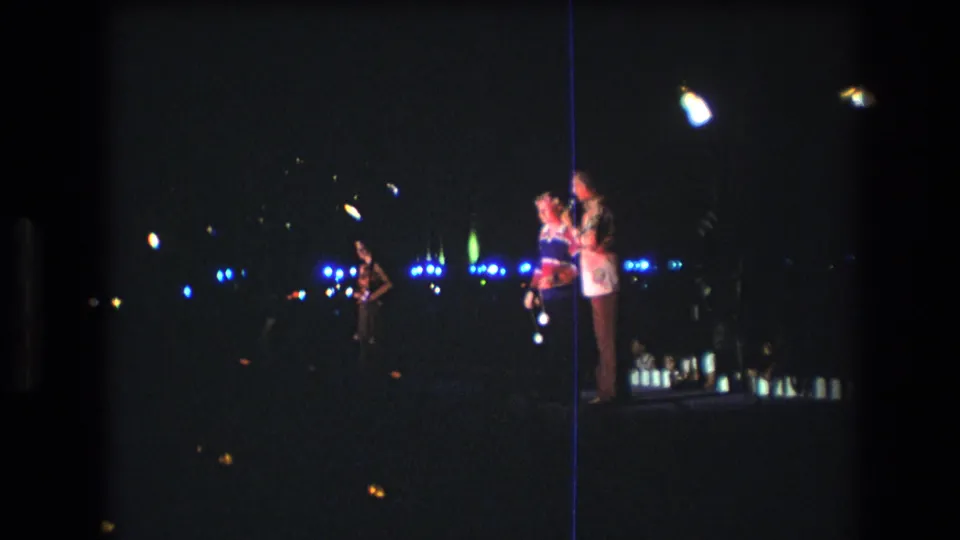 Low-light 1977 Honolulu home-movie frame with a couple standing together, another figure at left, and scattered blue lights across the dark background.