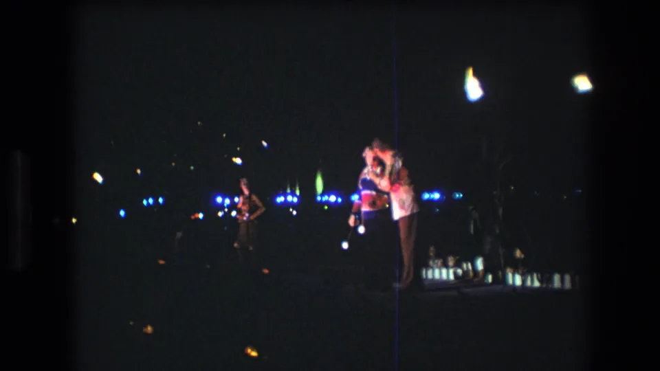 1977 Honolulu coastal night frame showing a couple in close contact, distant blue lights, bright flares, and visible 8mm film grain.