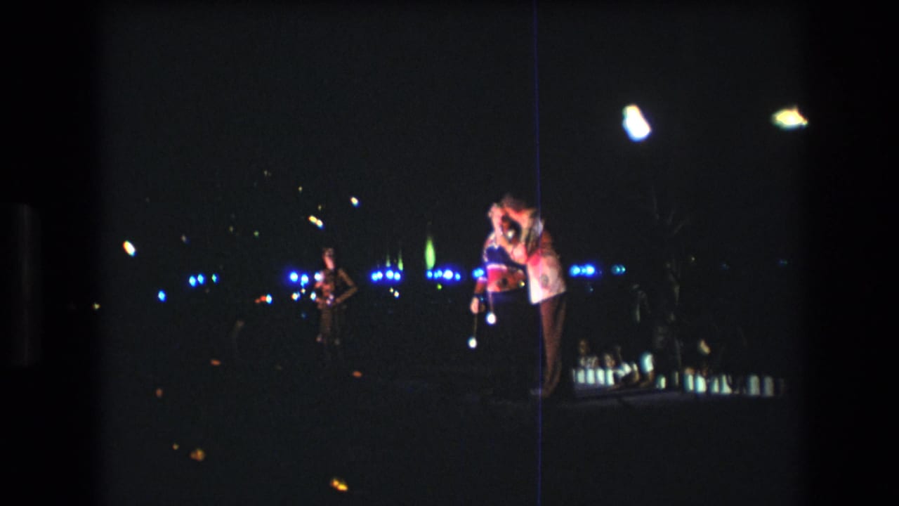 1977 Honolulu 8mm frame showing a dark stage or performance area with two people near a microphone, a musician at left, and blue-white lights in the background.