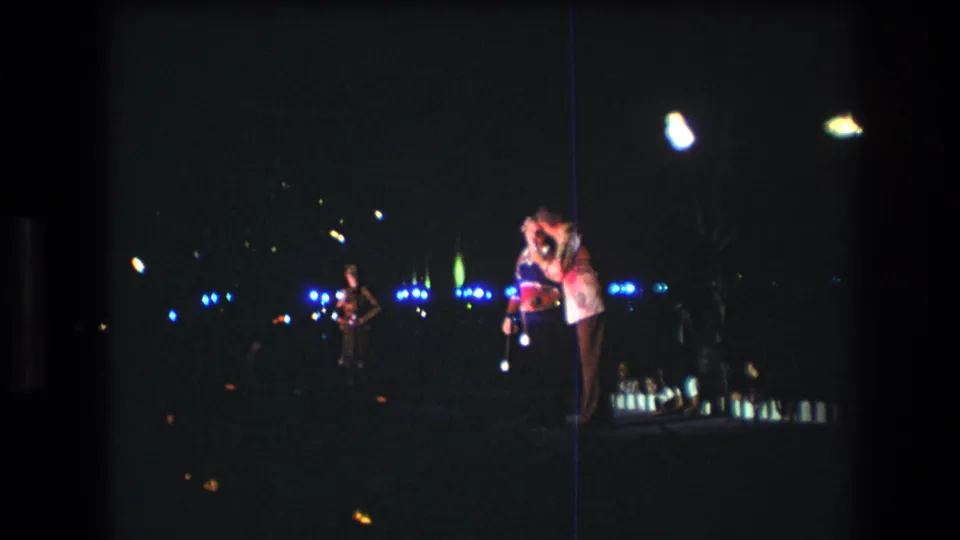 1977 Honolulu 8mm frame showing a dark stage or performance area with two people near a microphone, a musician at left, and blue-white lights in the background.