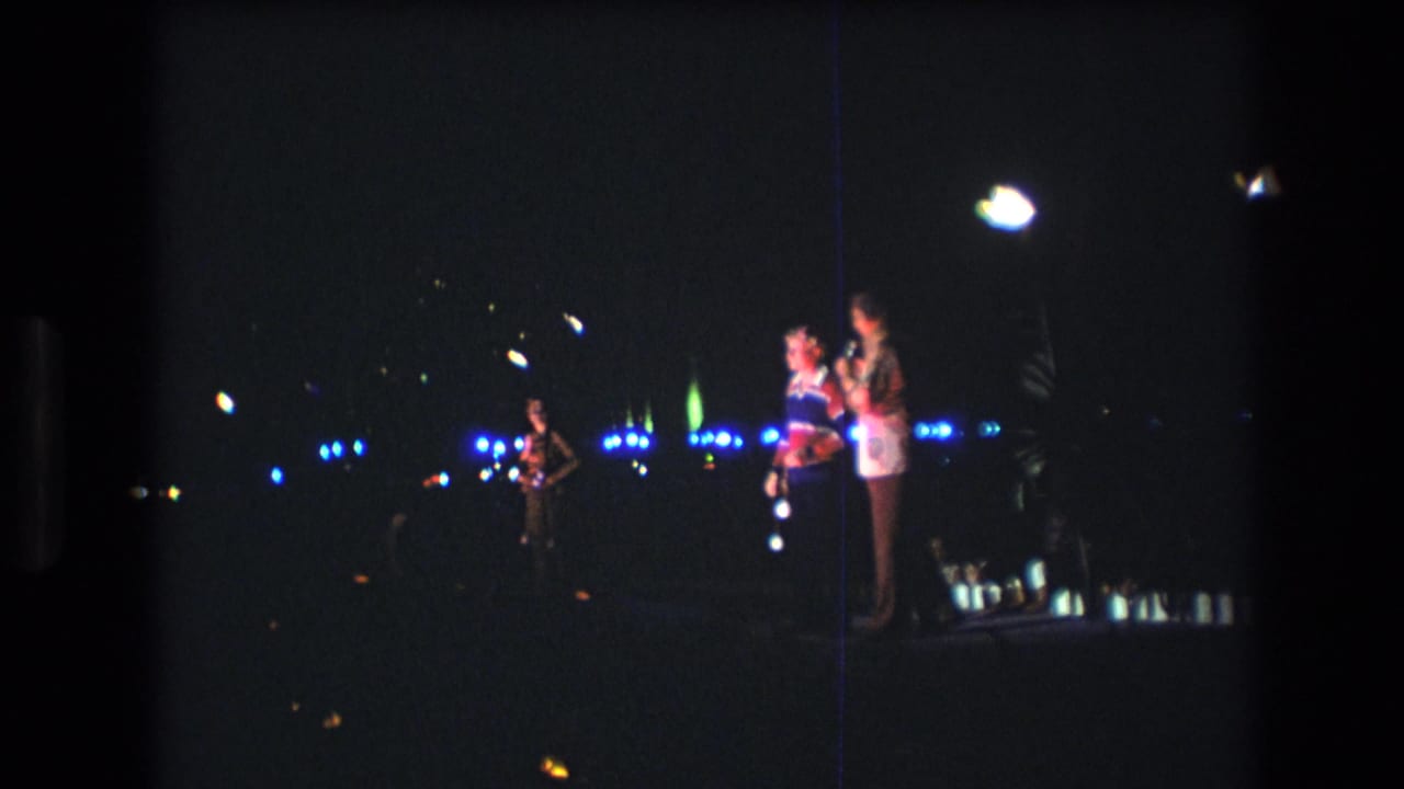 Two people stand on a dark stage while a musician performs at left.