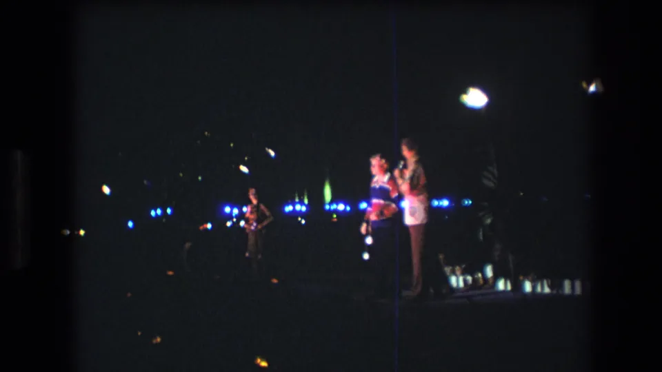 1977 Honolulu home-movie frame with two people on a dimly lit stage, a musician at left, and scattered blue and white lights across the dark background.