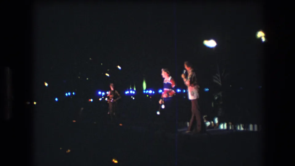 1977 Honolulu 8mm frame showing a person in a bright top near another performer, with a musician at left and colored lights behind them in a nighttime setting.