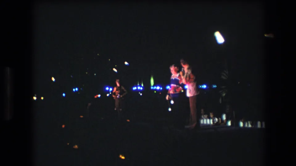1977 Honolulu home-movie frame of a nighttime stage performance with two figures near a microphone, a musician at left, and blue lights receding through the dark background.