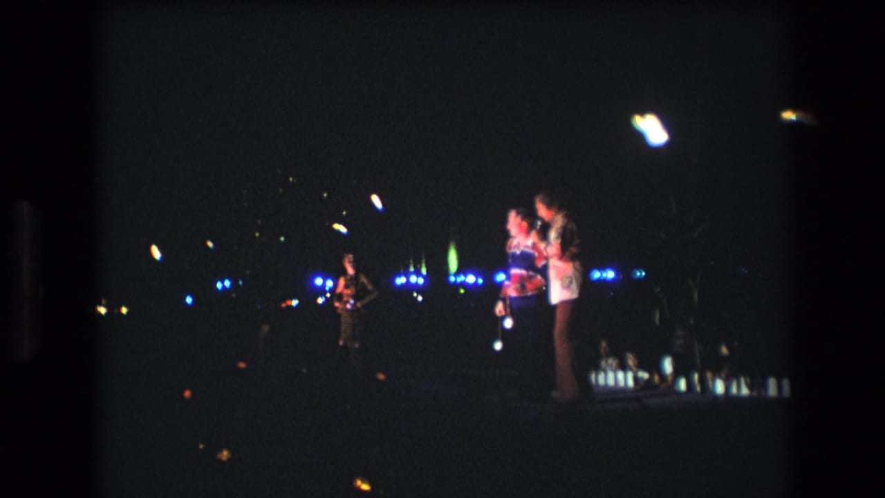1977 Honolulu 8mm frame showing two singers at microphones on a dim nighttime stage, with blue lights across the background and another performer at left.