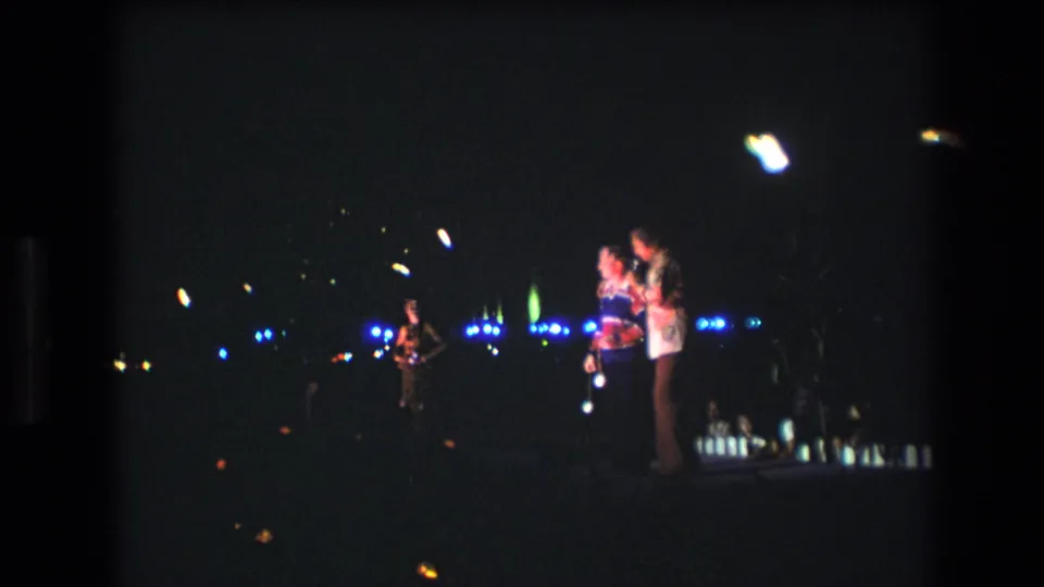 1977 Honolulu 8mm frame showing two singers at microphones on a dim nighttime stage, with blue lights across the background and another performer at left.