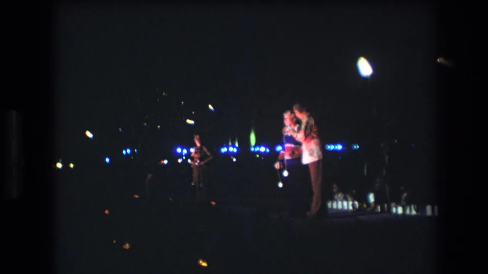 1977 Honolulu home-movie frame of a singing duet on a dark stage, with a bright overhead light, blue background lights, and a distant performer to the left.