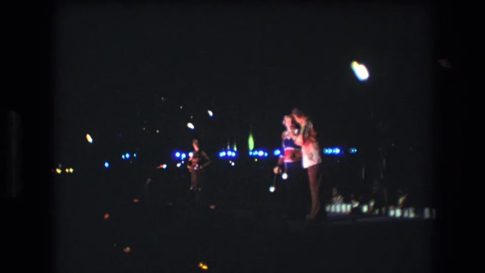 1977 Honolulu 8mm frame with a soft, dark view of two singers on stage, a performer at left, blue light points, and faint audience shapes below.