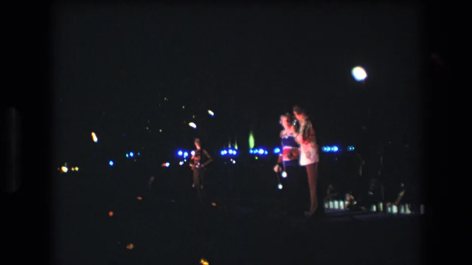 1977 Honolulu home-movie frame showing the duet near the right side of a nighttime stage, with blue decorative lights and faint audience presence at the lower edge.