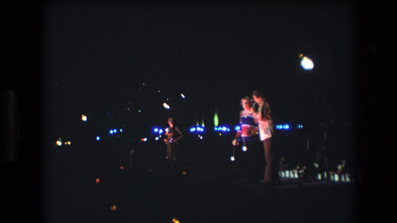 In 1977 Honolulu home-movie footage, a couple stands close together in a dark outdoor night scene with scattered blue and white lights behind them.