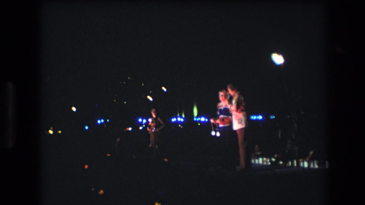 Two people stand together at night near a glowing object with scattered lights behind them.