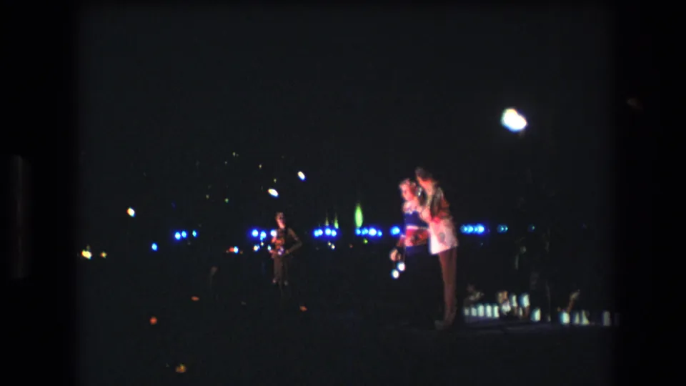 A couple remains close together in the low-light Honolulu footage, framed by grainy 8mm texture and scattered blue, green, and white light points.
