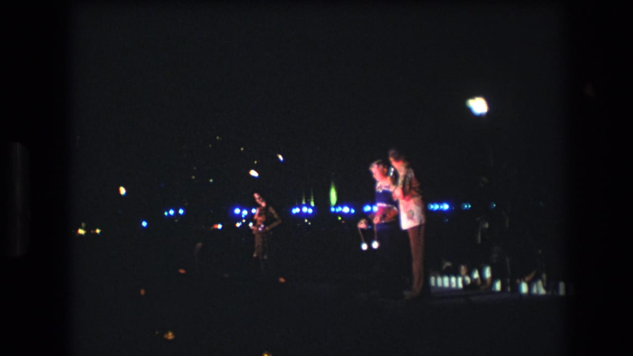 1977 Honolulu home-movie frame showing three people on a dim outdoor stage, with microphones, blue background lights, and a bright overhead flare.
