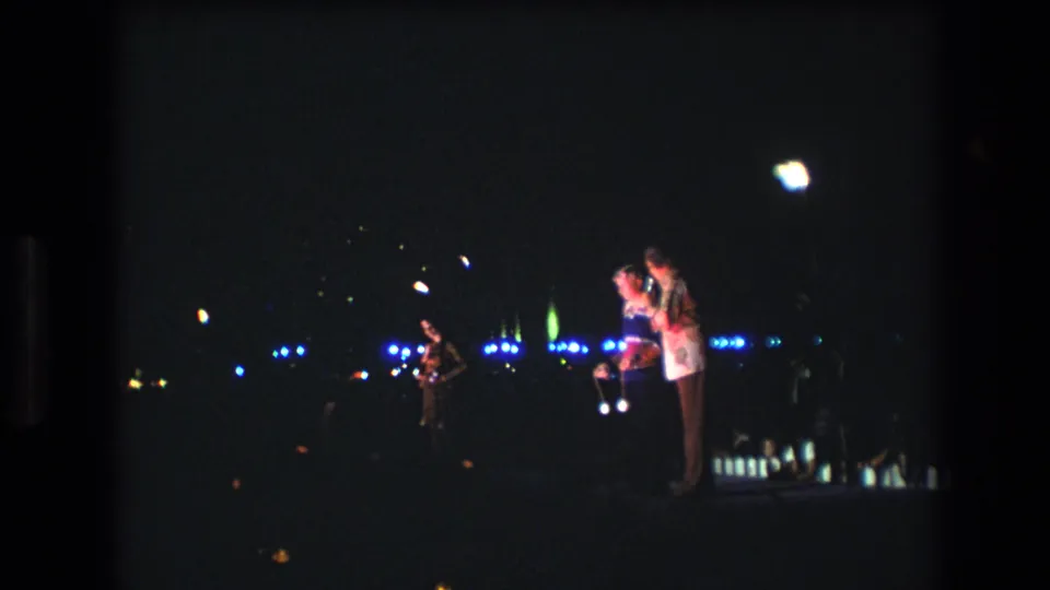 1977 Honolulu home-movie frame showing three people on a dim outdoor stage, with microphones, blue background lights, and a bright overhead flare.