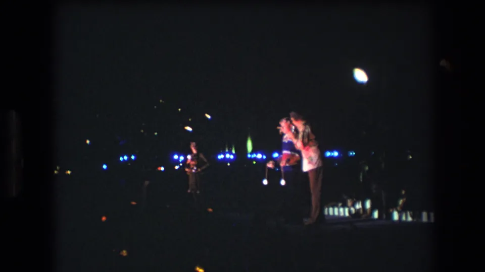 1977 Honolulu frame of a nighttime outdoor performance, with three microphone-holding figures on stage and scattered blue and green lights in the distance.