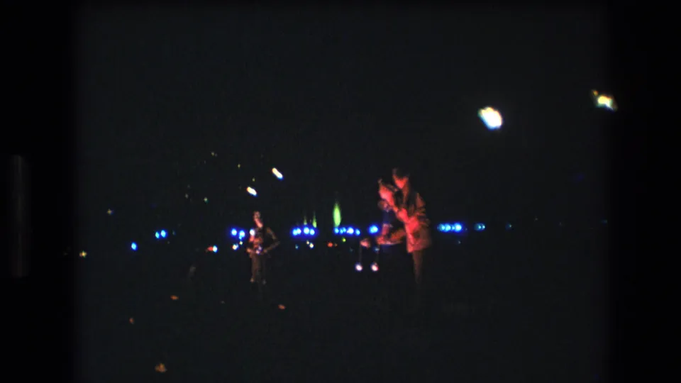 1977 Honolulu 8mm frame showing a shaky low-light view of three stage performers, microphone positions, blue light points, and strong film grain.