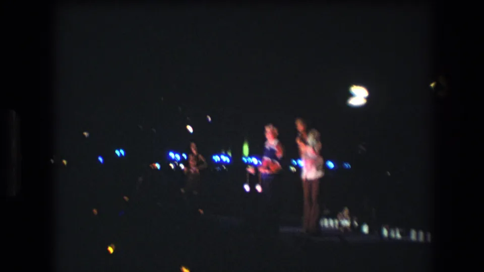 1977 Honolulu home-movie frame with three people clustered on a nighttime stage, blue background lights, overhead flares, and visible 8mm grain.