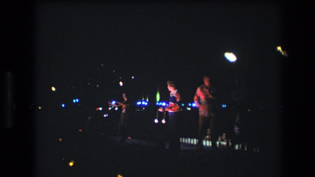 1977 Honolulu home-movie frame showing distant performers in a dark nighttime performance area with bright handheld or stage lights blooming in the frame.