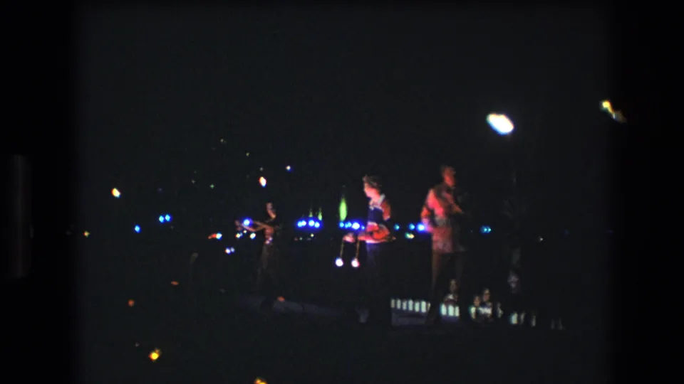 1977 Honolulu home-movie frame showing distant performers in a dark nighttime performance area with bright handheld or stage lights blooming in the frame.