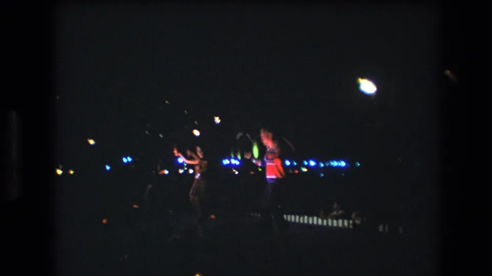 A dim 1977 Honolulu performance frame shows two distant figures against a dark background, with blue points of light and a bright overhead glow.
