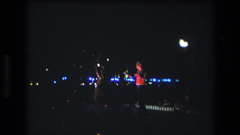 Low-light 8mm frame from Honolulu in 1977 showing a performer raising a glowing object while another figure stands nearby amid blue background lights.