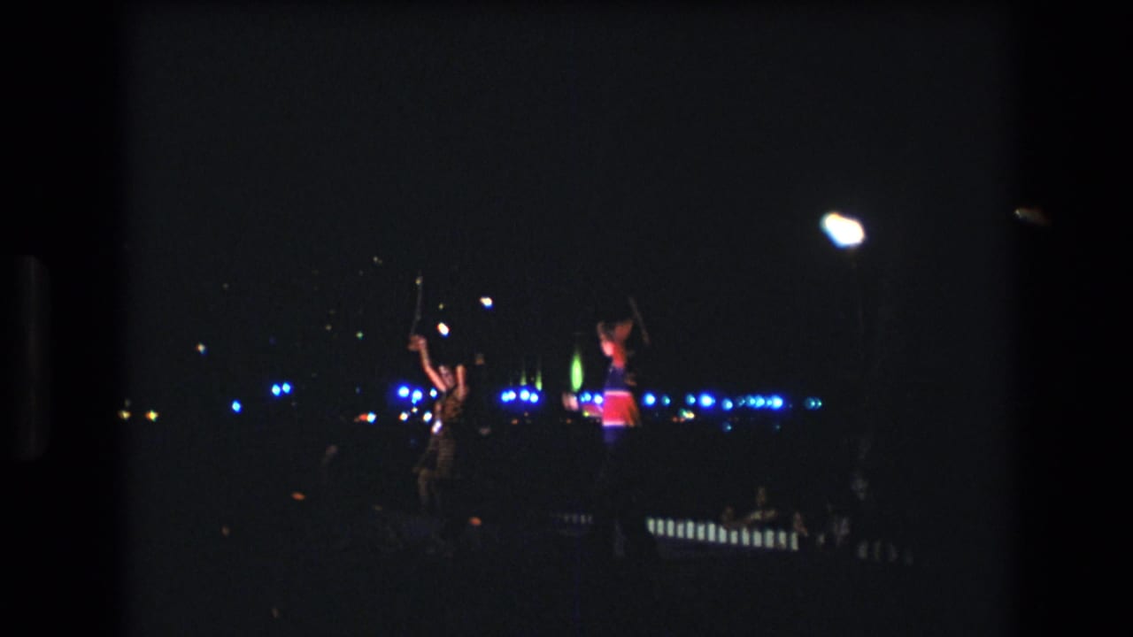 In this 1977 Honolulu, Oahu home-movie frame, two distant performers move on a dim nighttime stage beneath bright white lamps and blue background lights.