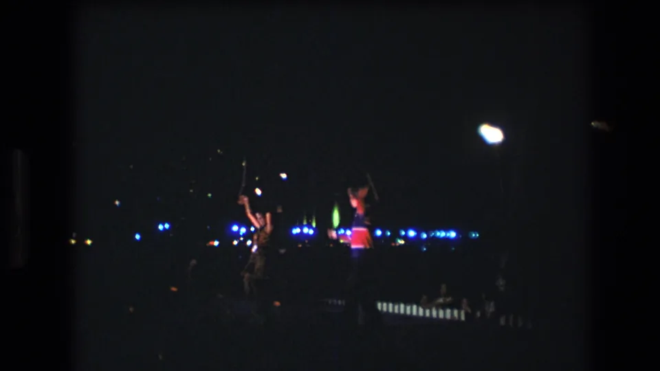 In this 1977 Honolulu, Oahu home-movie frame, two distant performers move on a dim nighttime stage beneath bright white lamps and blue background lights.