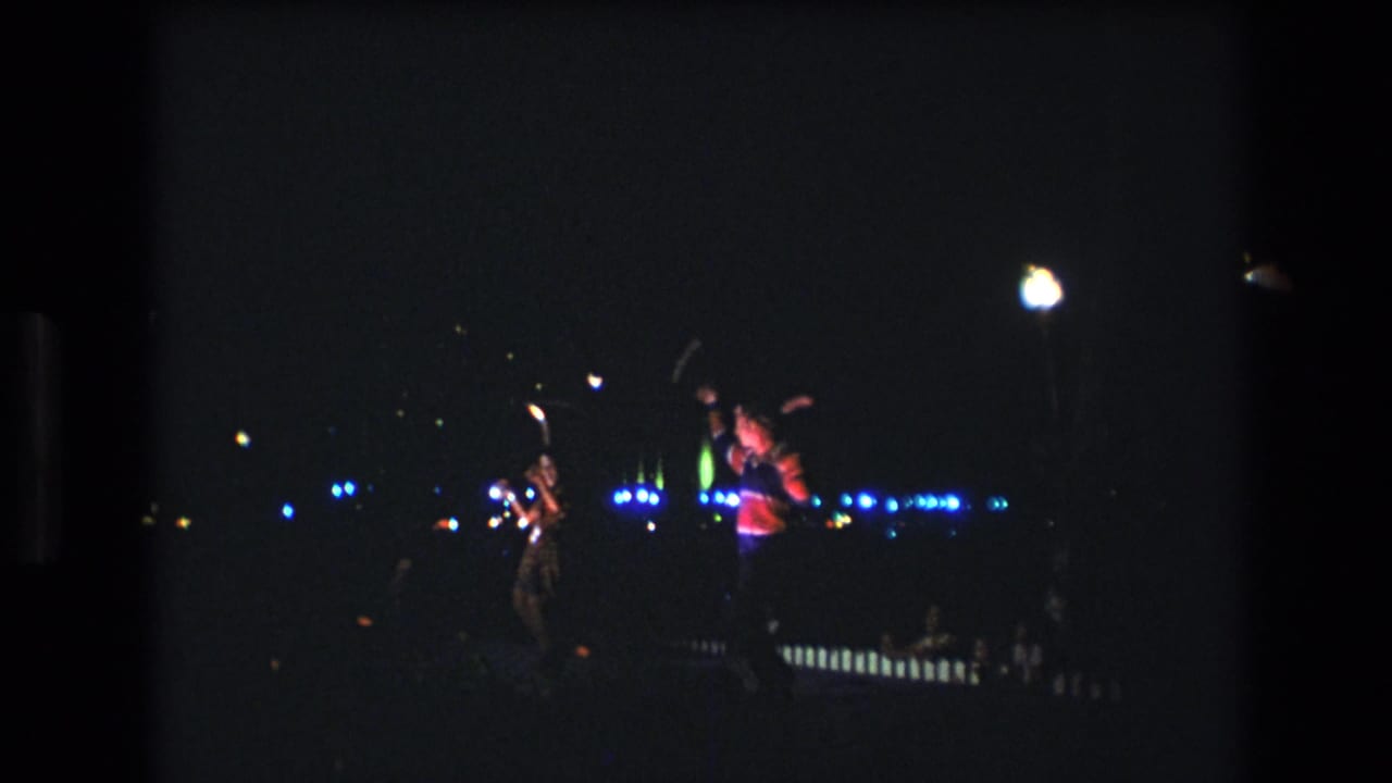 Performers move across a dark performance area with bright lamps and blue lights in the distance.