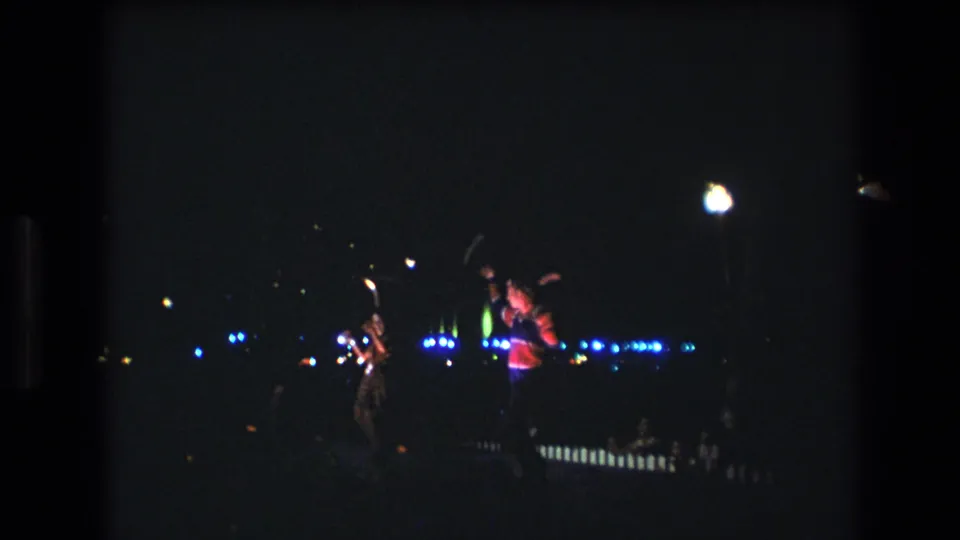 The frame shows a low-light 1977 Honolulu stage performance with two distant figures, one in a red top, framed by scattered blue and white lights.