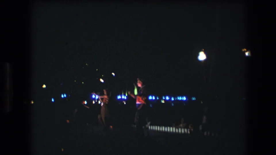 This 1977 Honolulu, Oahu frame captures two performers in a dark nighttime setting, with blue light points across the background and a bright overhead lamp.