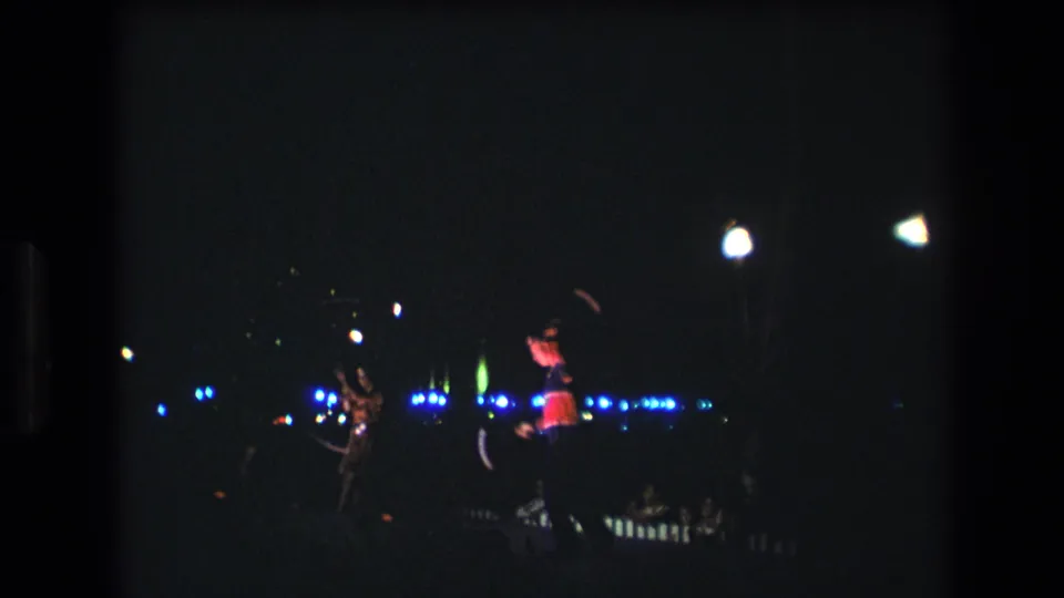 A dark 8mm home-movie frame from 1977 Honolulu shows two distant stage performers, one partly lit in red, against scattered blue and white nighttime lights.
