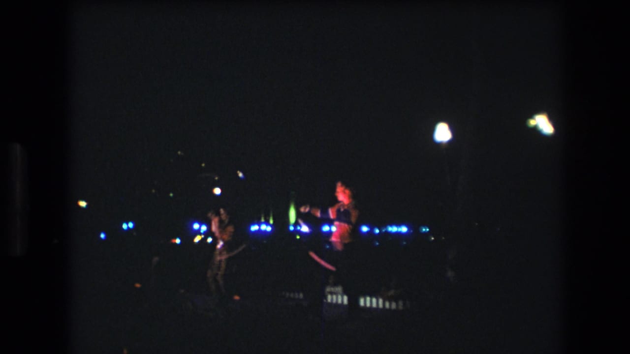 Two women in sparkling costumes appear to twirl batons during a nighttime 1977 Honolulu performance, seen from a distant Super 8 home-movie viewpoint.