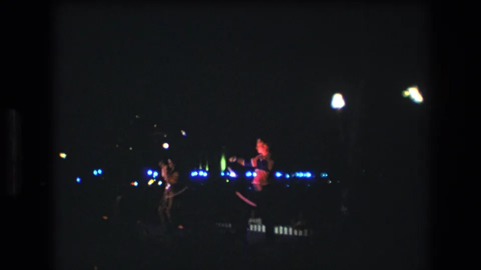 Two women in sparkling costumes appear to twirl batons during a nighttime 1977 Honolulu performance, seen from a distant Super 8 home-movie viewpoint.