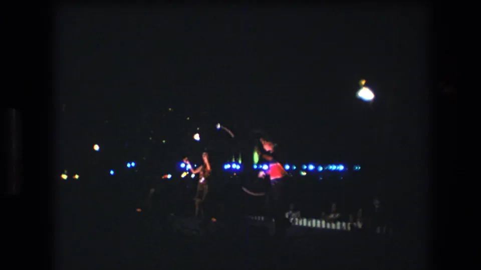 Two baton performers stand apart in a dark Honolulu nighttime setting, with bright blue lights and small white highlights visible behind them.