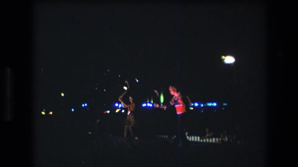 The low-light Super 8 frame shows two women continuing a baton routine at night, with a bright overhead light and blue background points across the dark scene.