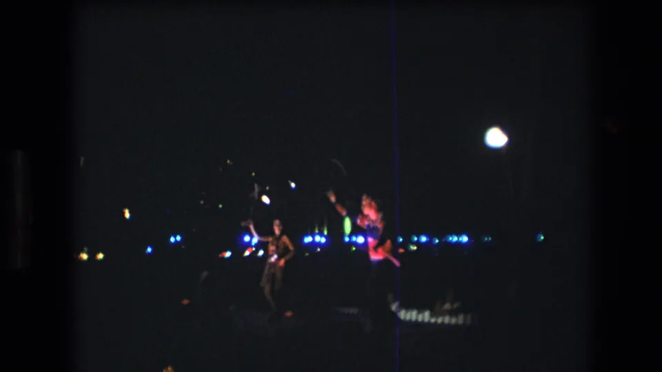 One woman raises a baton or arm while another performer stands nearby during the nighttime Honolulu routine, with blue lights stretching across the background.