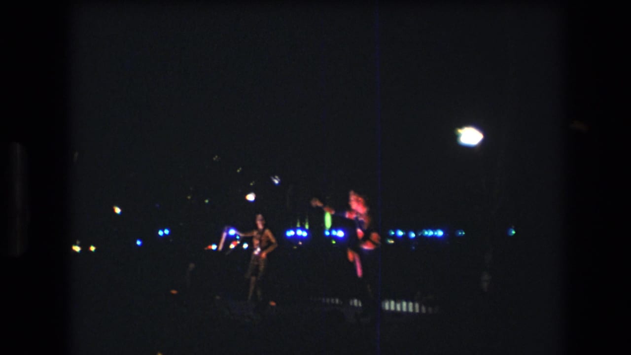 1977 Honolulu-area 8mm home-movie frame showing two distant nighttime performers with glowing props against blue festival lights.