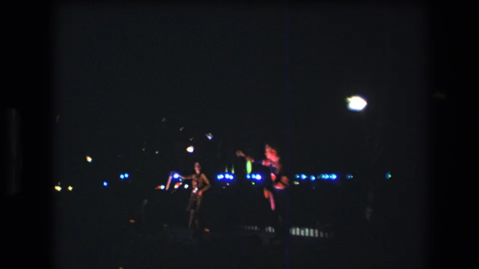 1977 Honolulu-area 8mm home-movie frame showing two distant nighttime performers with glowing props against blue festival lights.