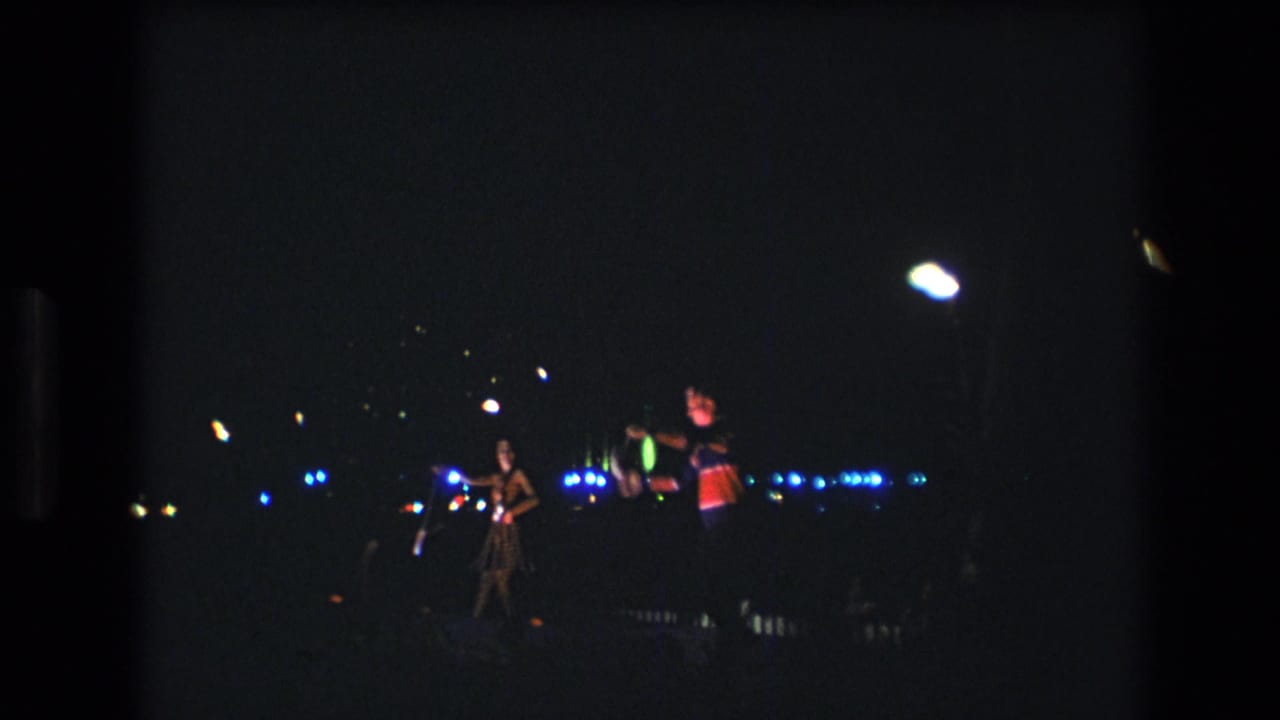 A low-light 1977 Honolulu 8mm frame with performers silhouetted near blue lights and scattered glowing props.