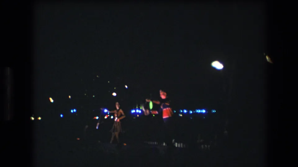 A low-light 1977 Honolulu 8mm frame with performers silhouetted near blue lights and scattered glowing props.
