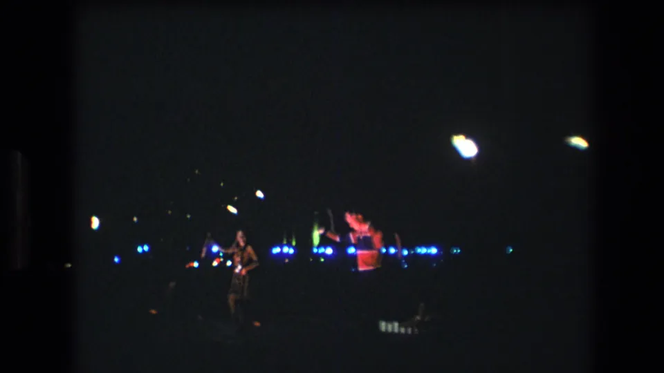 1977 Honolulu home-movie frame showing a distant performer in a patterned outfit near glowing props and blue nighttime lights.