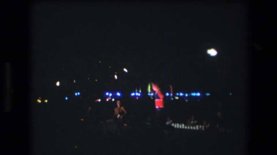 A 1977 Honolulu 8mm night frame with a performer raising glowing props near a row of blue festival lights.