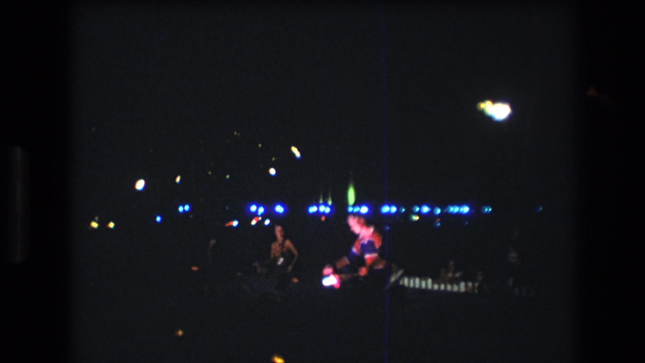 1977 Honolulu 8mm footage shows distant performers on a dark stage or performance area under blue lights, with heavy grain and a vertical scratch visible.