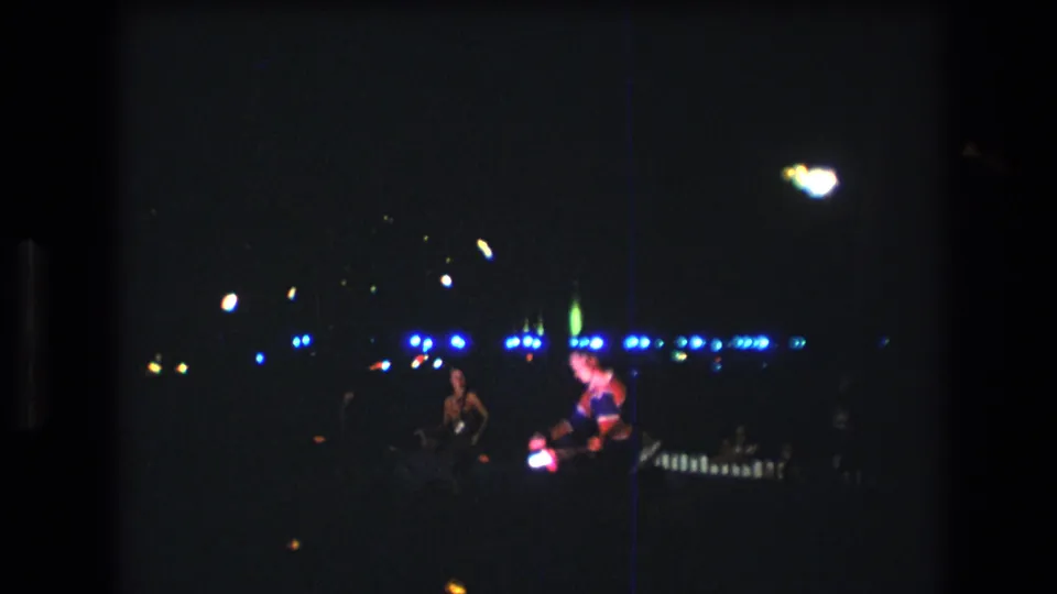 1977 Honolulu 8mm footage shows distant performers on a dark stage or performance area under blue lights, with heavy grain and a vertical scratch visible.