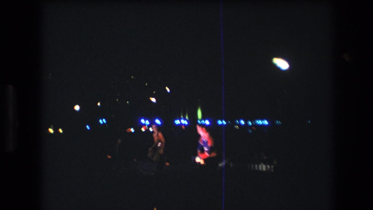 Two distant performers appear near blue lights on a dark stage.