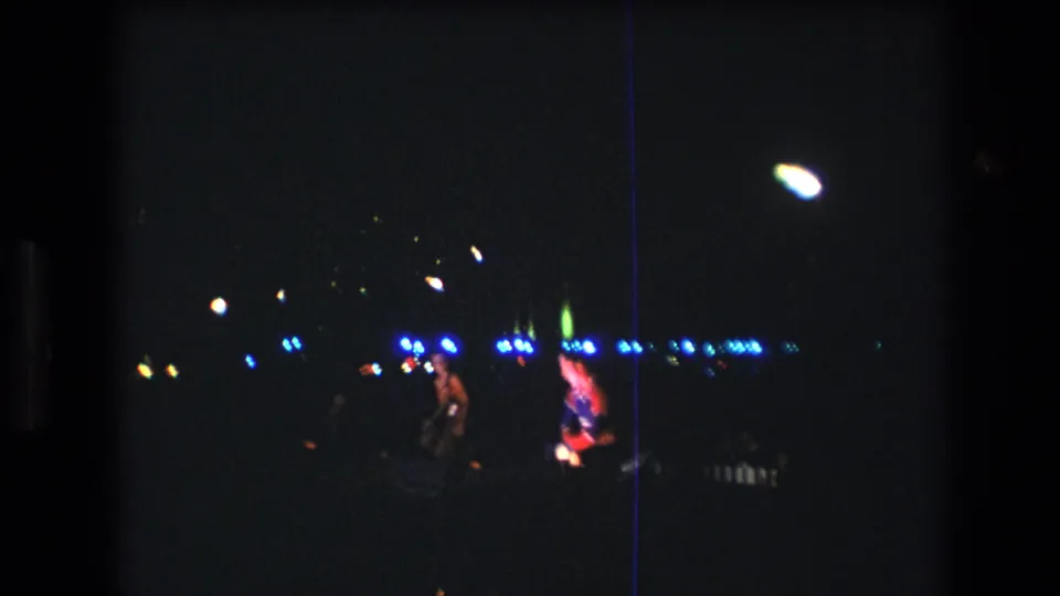 A dim 1977 Honolulu stage scene shows performers in traditional-style attire moving near blue lights, filmed from a distant audience viewpoint on 8mm.